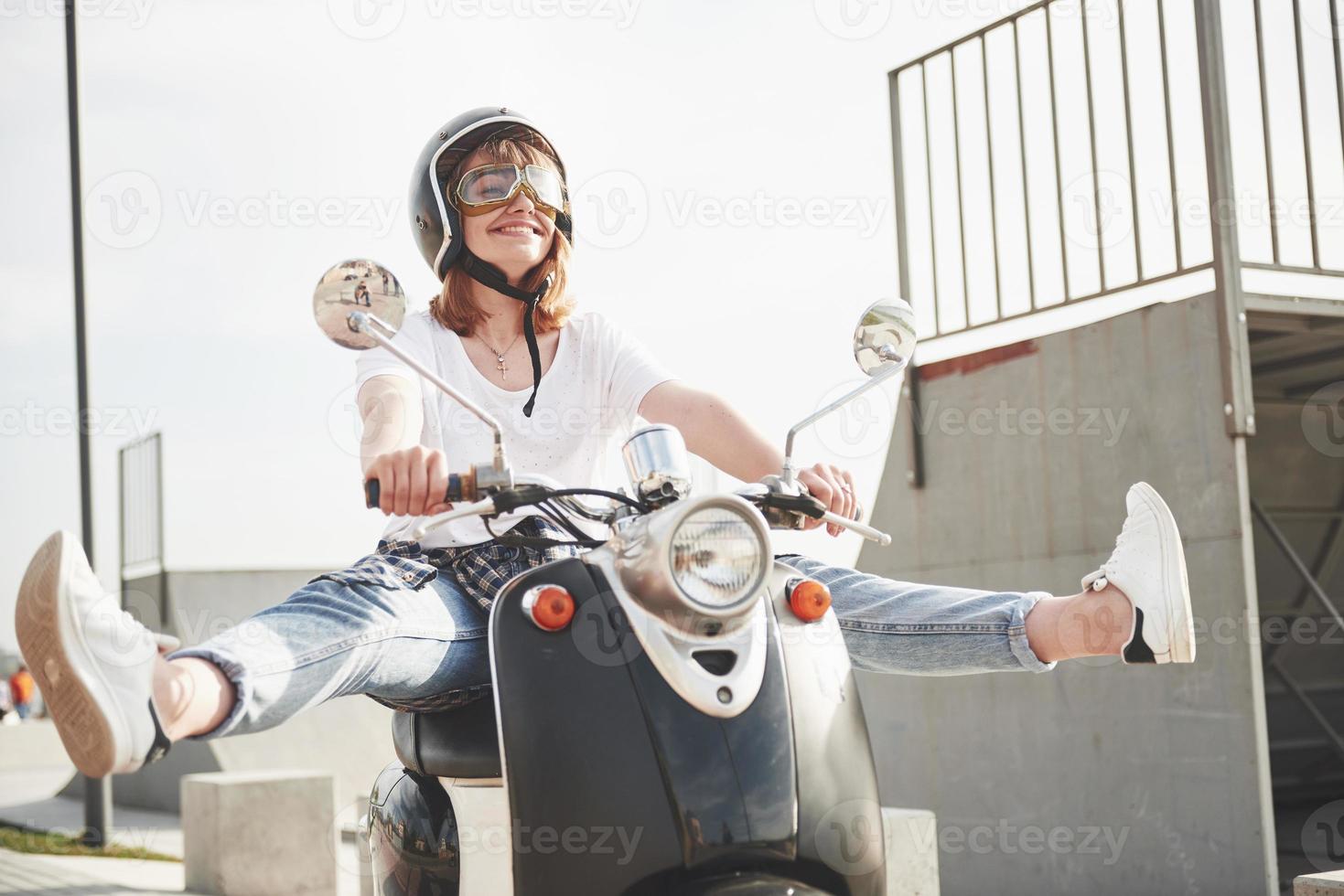 retrato de um hipster linda garota sentada em uma scooter retrô preta, sorrindo, posando e desfrutar do sol quente de primavera. foto