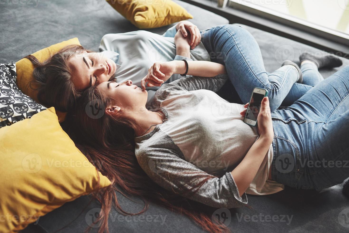 duas irmãs gêmeas sorridentes fofos segurando o smartphone e fazendo selfie. garotas deitam no sofá posando e alegria foto