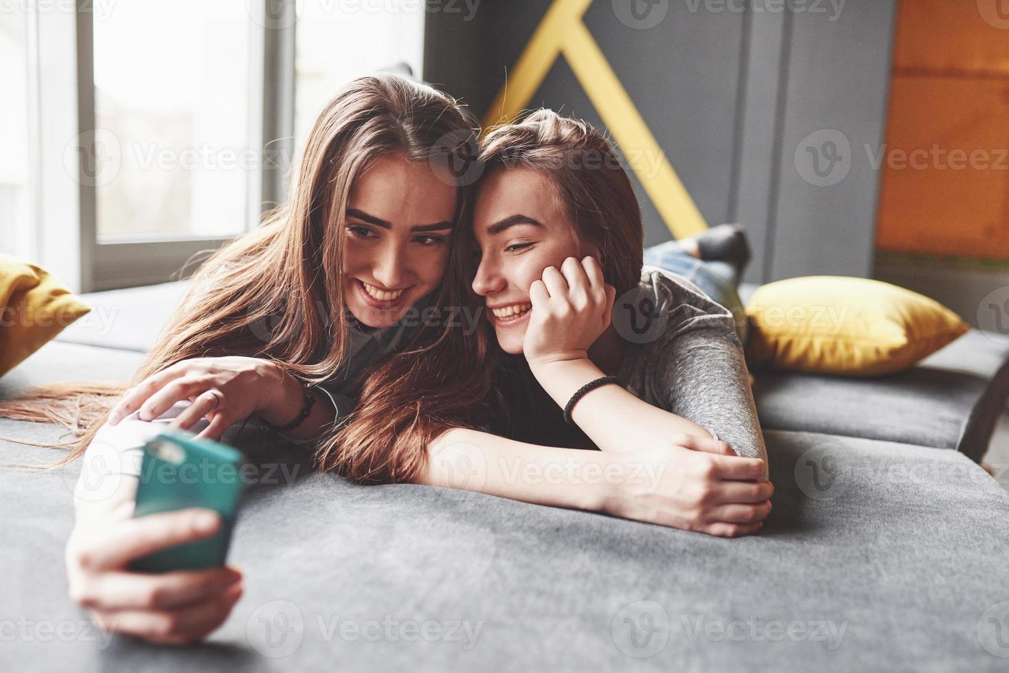 duas irmãs gêmeas sorridentes fofos segurando o smartphone e fazendo selfie. garotas deitam no sofá posando e alegria foto