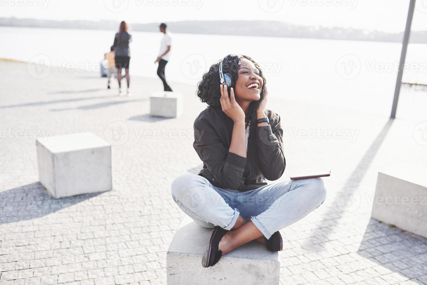 retrato de uma bela jovem afro-americana sentada na praia ou lago e ouvindo música em seus fones de ouvido foto