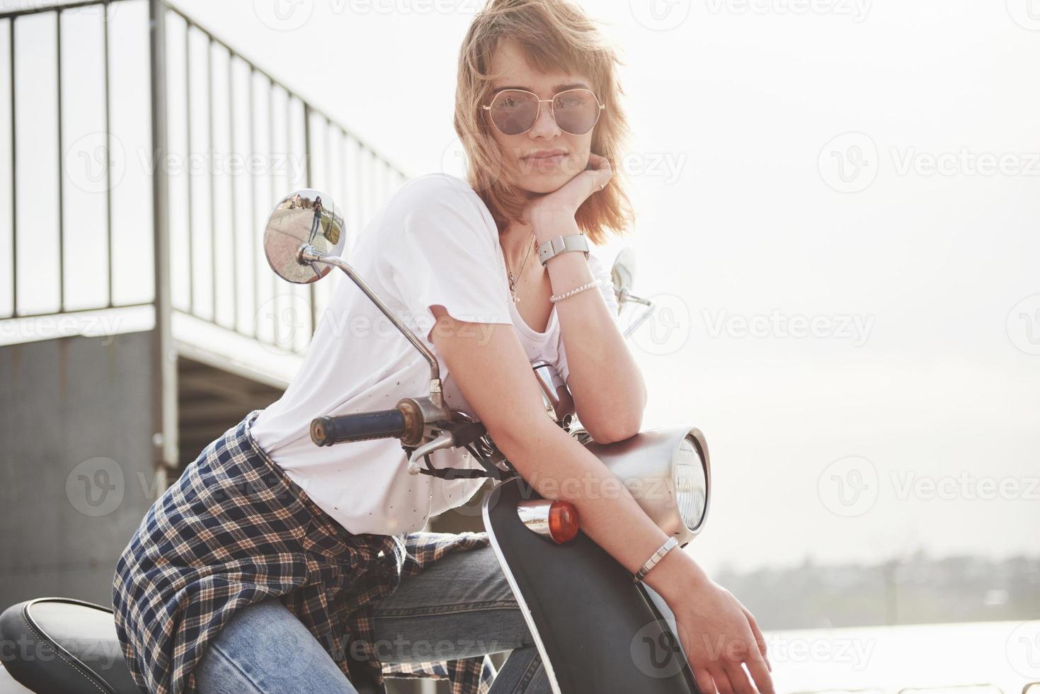retrato de um hipster linda garota sentada em uma scooter retrô preta, sorrindo, posando e desfrutar do sol quente de primavera. foto