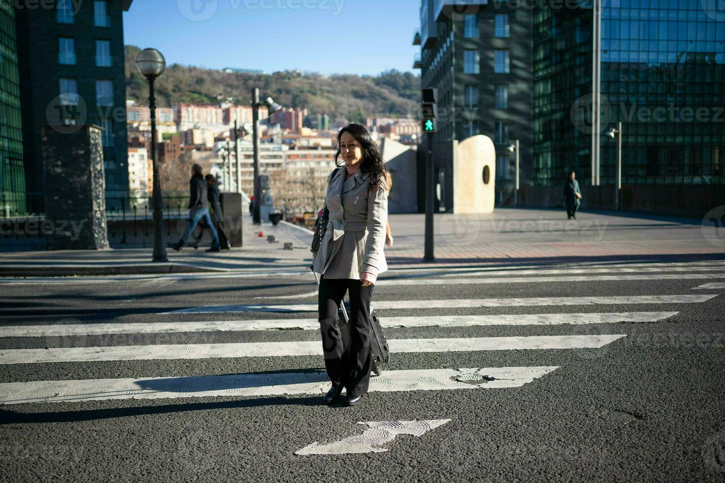 morena executivo mulher dentro uma cinzento casaco, cruzando uma estrada com dela trabalhos mala. foto