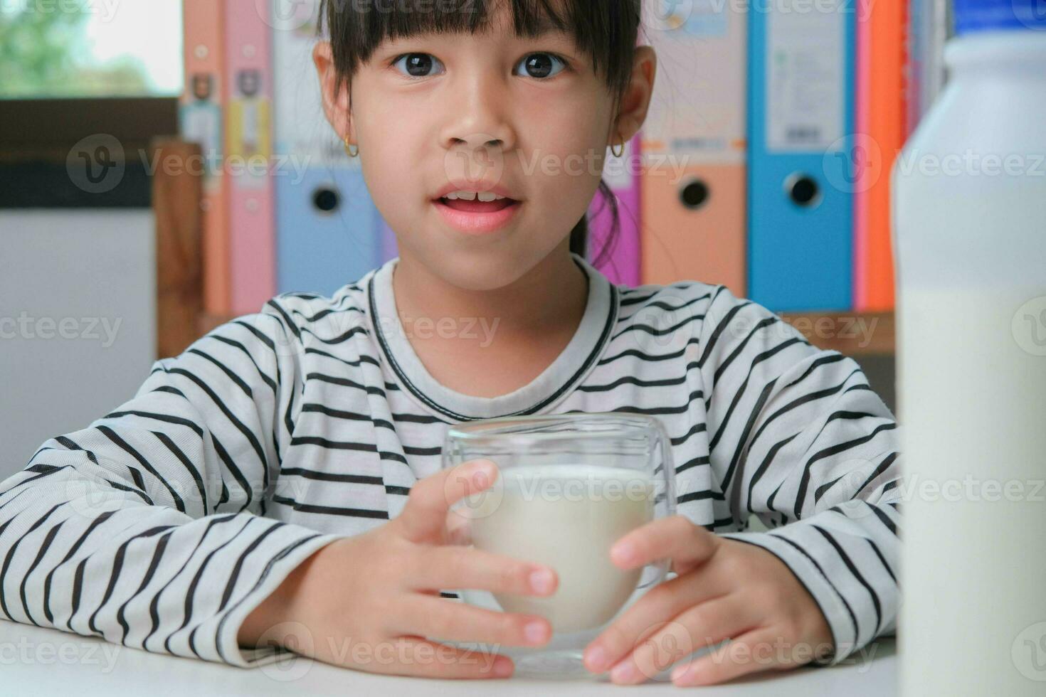 fofa ásia menina bebendo uma vidro do leite às casa dentro vivo sala. pequeno menina bebendo leite dentro a manhã antes indo para escola. saudável Comida dentro infância. foto