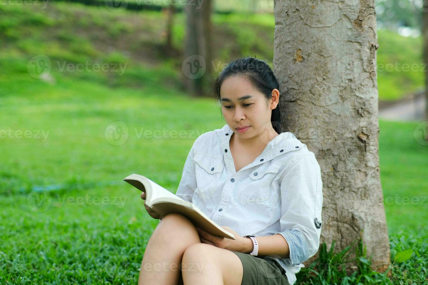 alegre jovem mulher dentro Jaqueta lendo uma livro dentro verão parque. concentrado mulher sentado em Relva e estudando debaixo árvore durante período de férias. Educação conceito foto