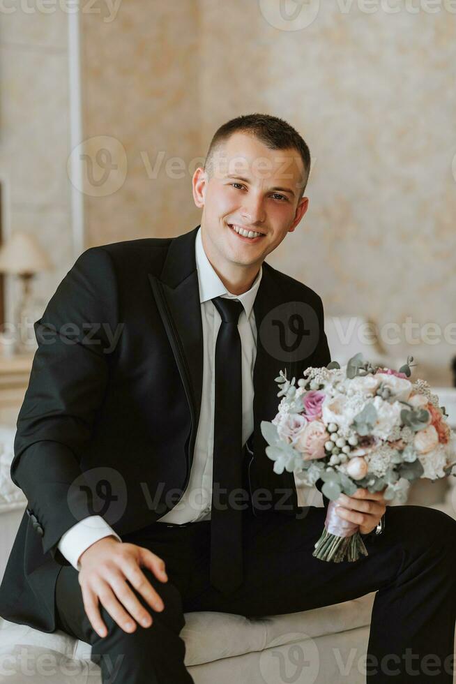 jovem homem sentado dentro uma cadeira com uma ramalhete do flores dentro uma hotel sala, uma jovem homem de negocios dentro uma Preto terno e branco camisa com uma gravata. a noivo é preparando para a Casamento cerimônia. foto