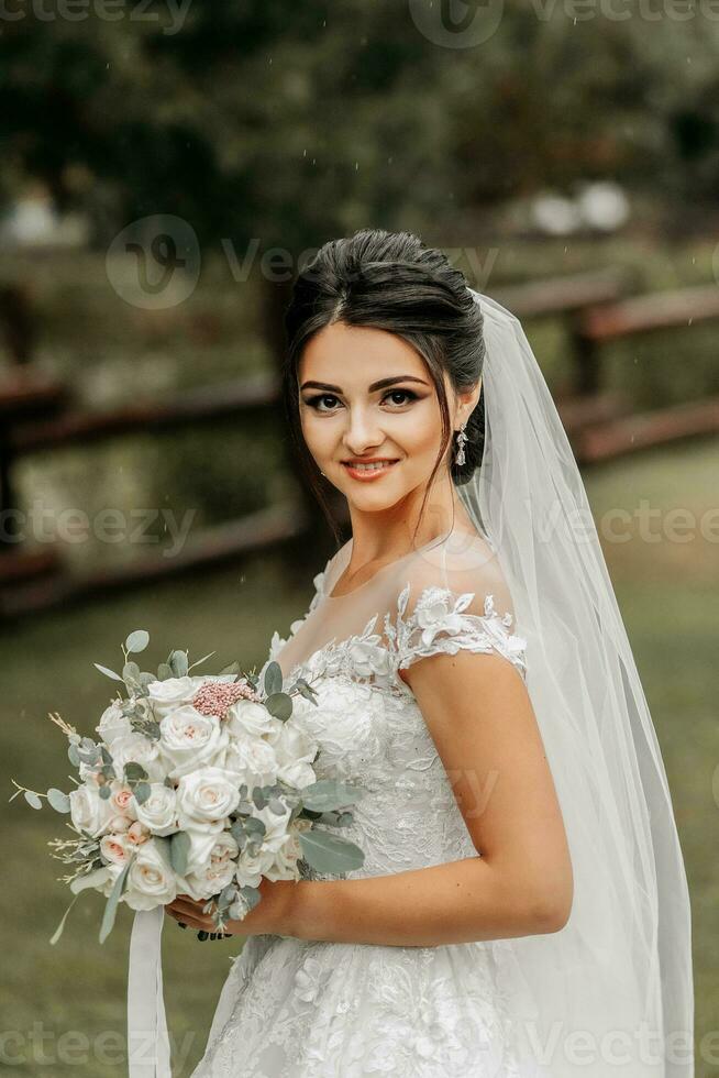 retrato do uma morena noiva dentro uma branco Casamento vestir com uma Casamento ramalhete dentro a parque. cheio comprimento foto. branco renda Casamento vestir foto