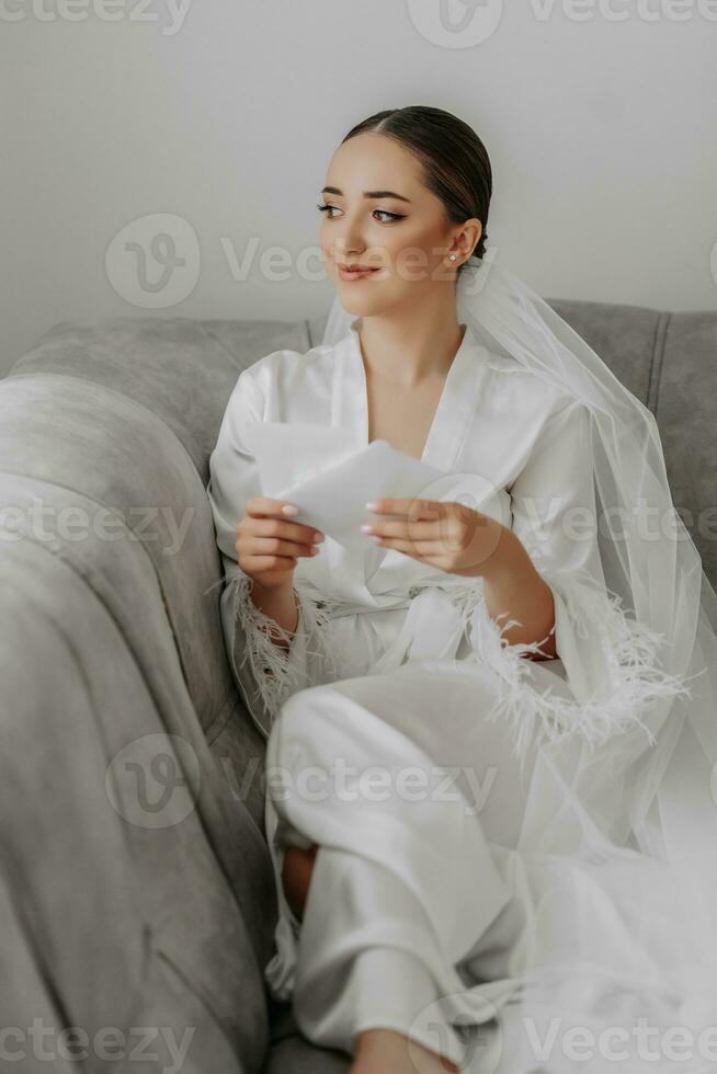 retrato do lindo menina noiva com à moda penteado e Maquiagem dentro branco roupão sentado em sofá e segurando Casamento convite. foto