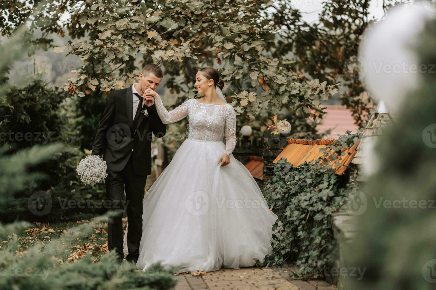 uma à moda casal do europeu recém-casados. sorridente noiva dentro uma branco vestir. a noivo é vestido dentro uma clássico Preto terno, branco camisa e gravata. Casamento dentro natureza, uma andar dentro a jardim entre alta árvores foto