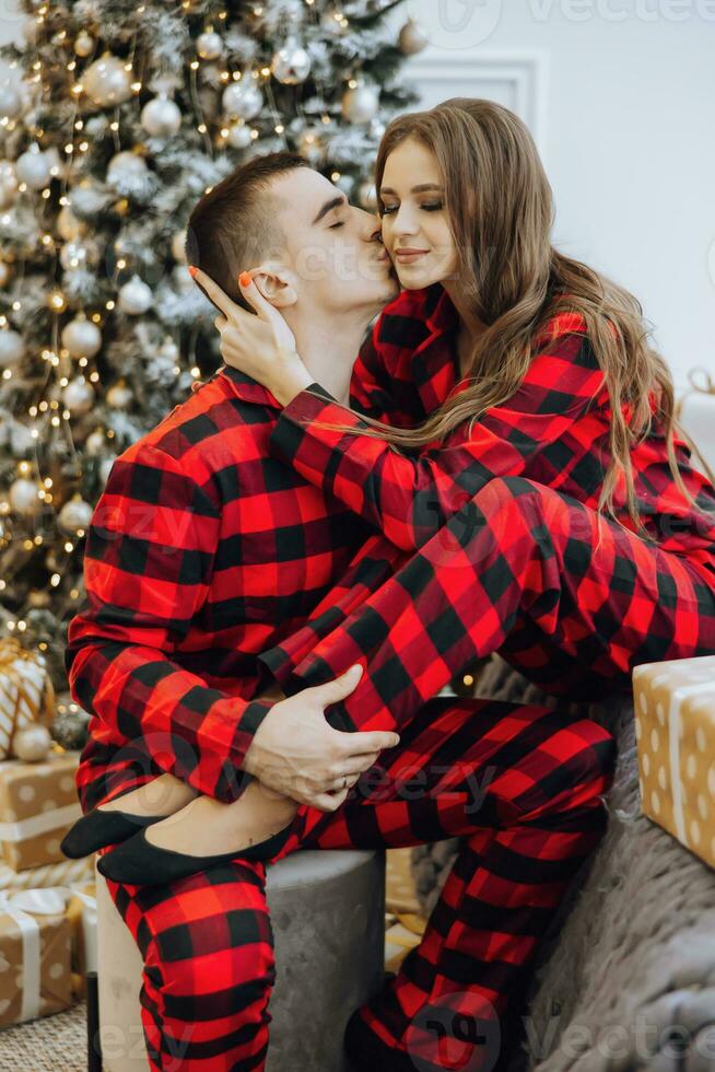 uma jovem lindo casado casal dentro a mesmo vermelho pijamas, perto a Natal árvore, estão sorridente, abraçando e se beijando. Novo ano feriados e presentes debaixo a Natal árvore foto
