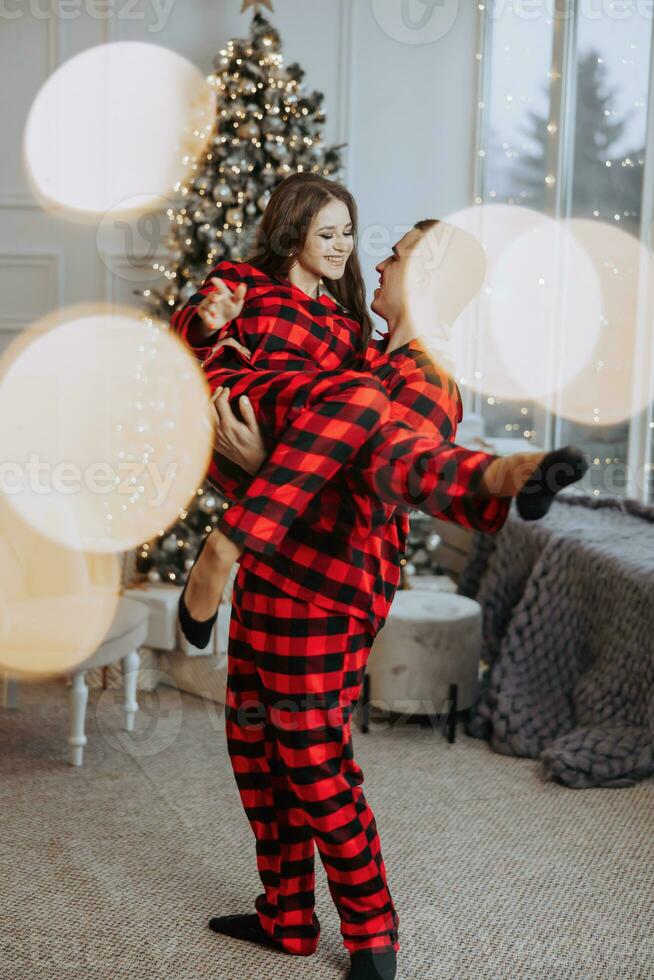 uma lindo jovem menina dentro vermelho pijamas carrinhos em a peitoril da janela e coloca em uma Natal árvore. Natal conceito. muitos presentes dentro papel invólucros debaixo a Natal árvore foto