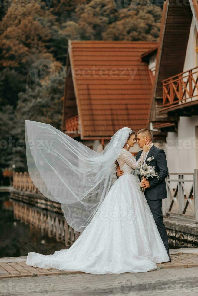 Casamento retrato. uma morena noiva dentro uma grandes vestir e uma noivo dentro uma clássico terno estão de pé, abraçando dentro amor contra a fundo do uma lago e uma castelo. a véu é dentro a ar foto