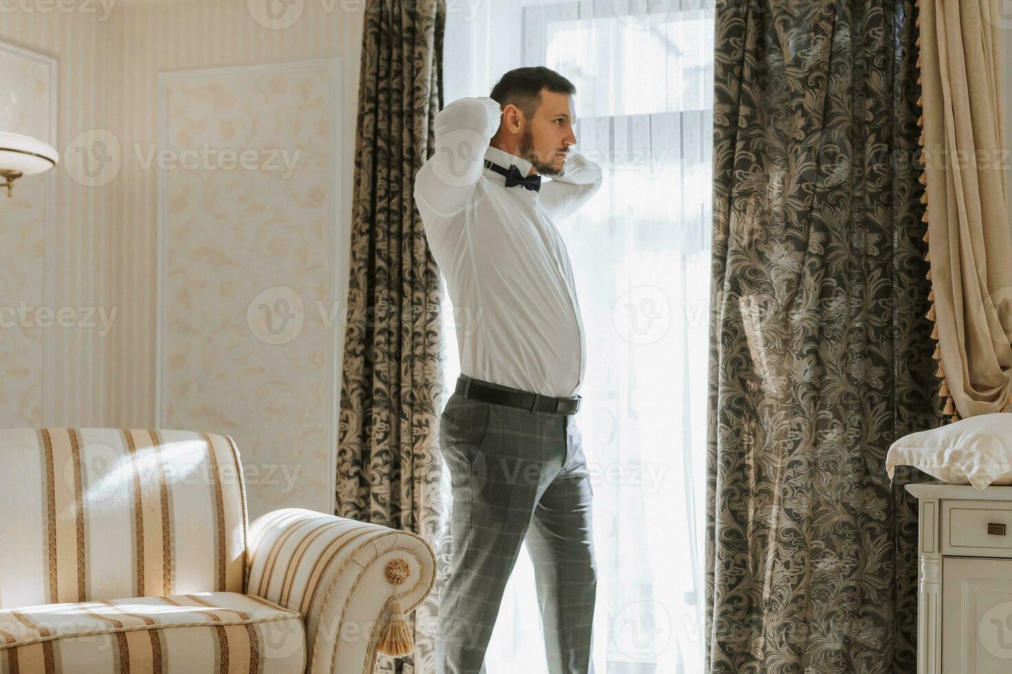 uma à moda noivo com uma barba é preparando para a Casamento cerimônia. do noivo manhã. uma homem de negocios aperta uma Preto arco gravata. a noivo é obtendo pronto dentro a manhã antes a Casamento cerimônia foto