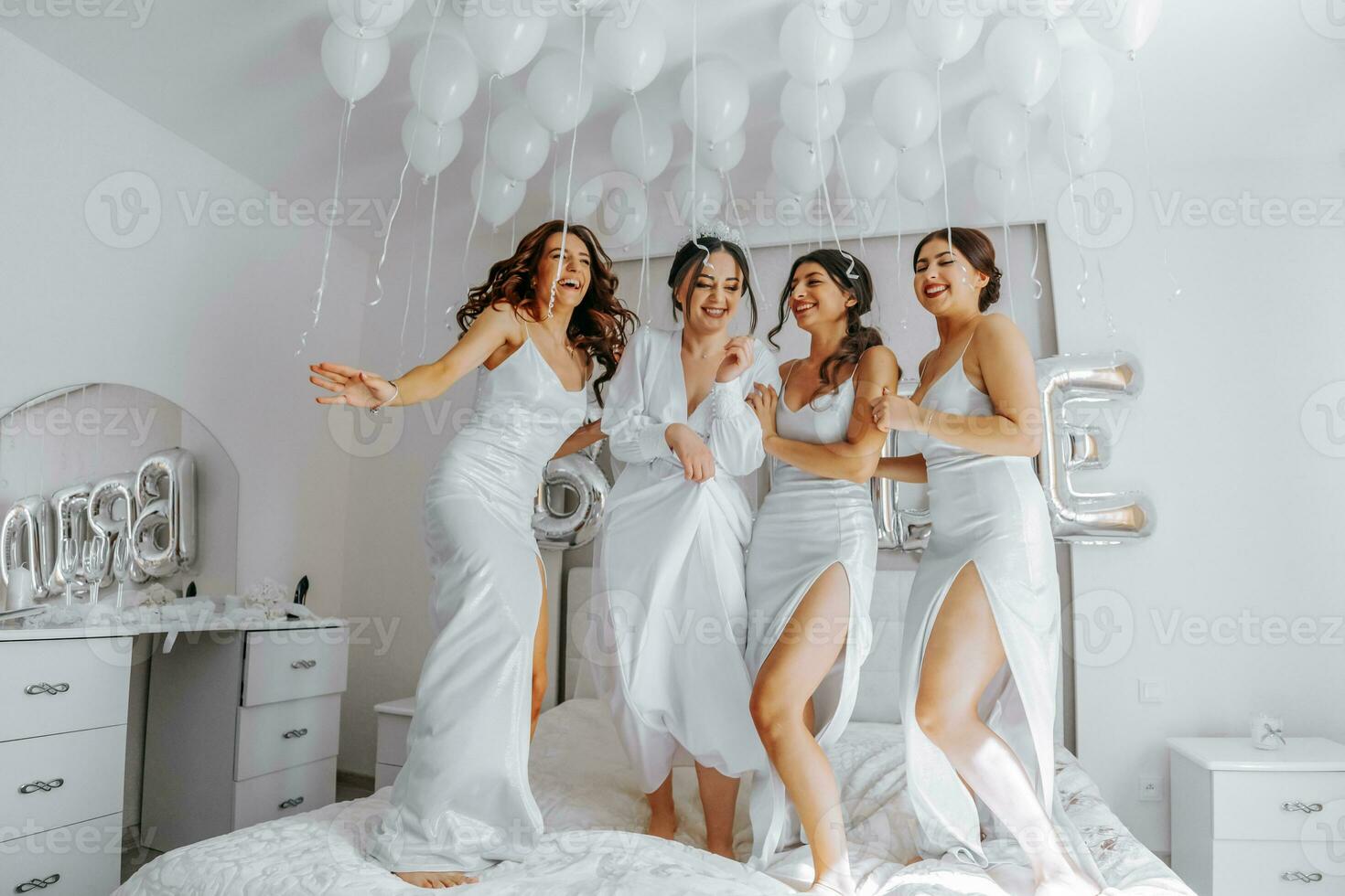 jovem damas de honra dentro branco seda vestidos em uma cama dentro a da noiva sala. lindo mulheres a comemorar solteira festa sentado em cama. foto