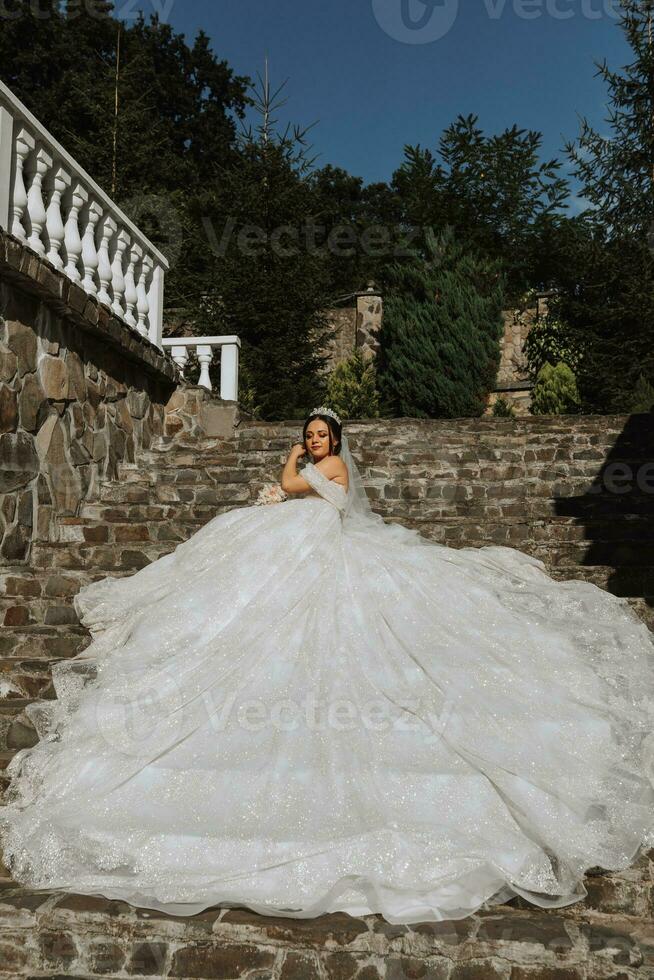 jovem lindo noiva dentro uma Casamento vestir com aberto ombros e uma coroa em dela cabeça é deitado em pedra escadaria, moda tiro debaixo severo luz solar foto
