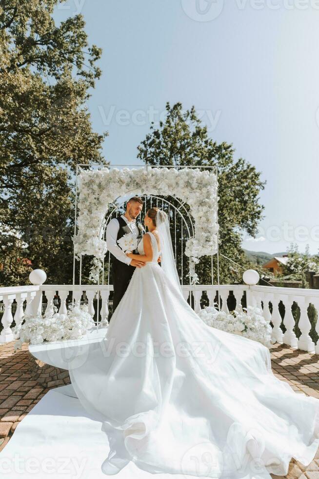 à moda noivo dentro branco camisa e Preto colete e fofa Loiras noiva dentro branco vestir dentro parque perto elegante branco rosa arco. Casamento retrato do recém-casados. foto