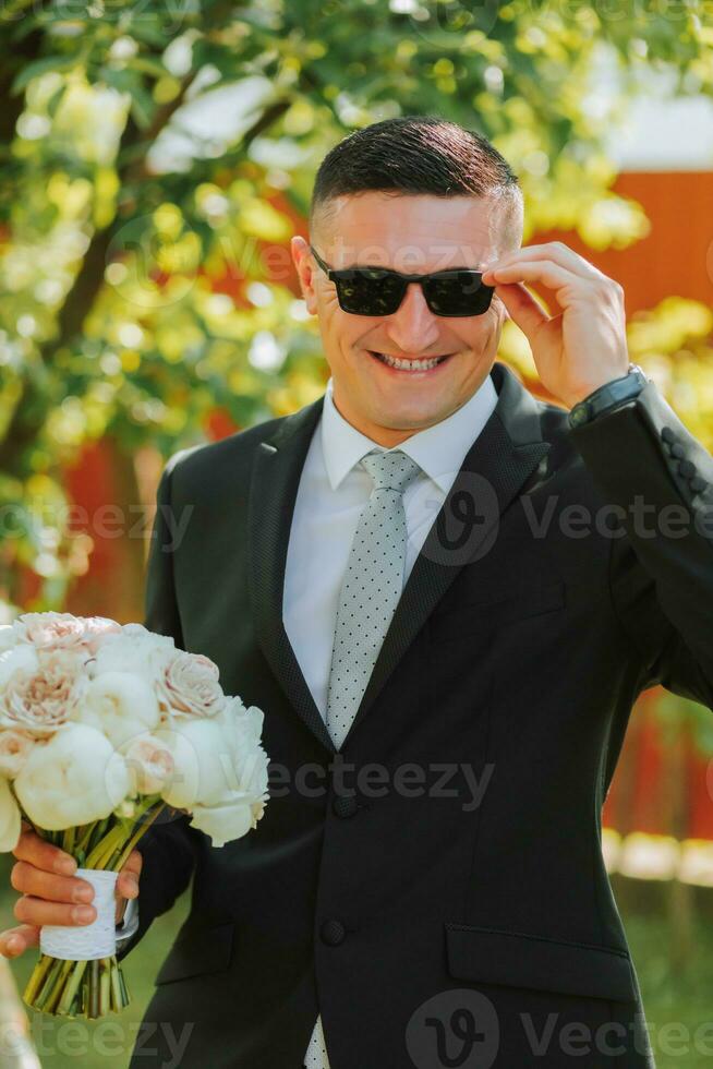 foto do a noivo dentro uma clássico Preto terno e copos, com uma ramalhete do flores dentro dele mãos