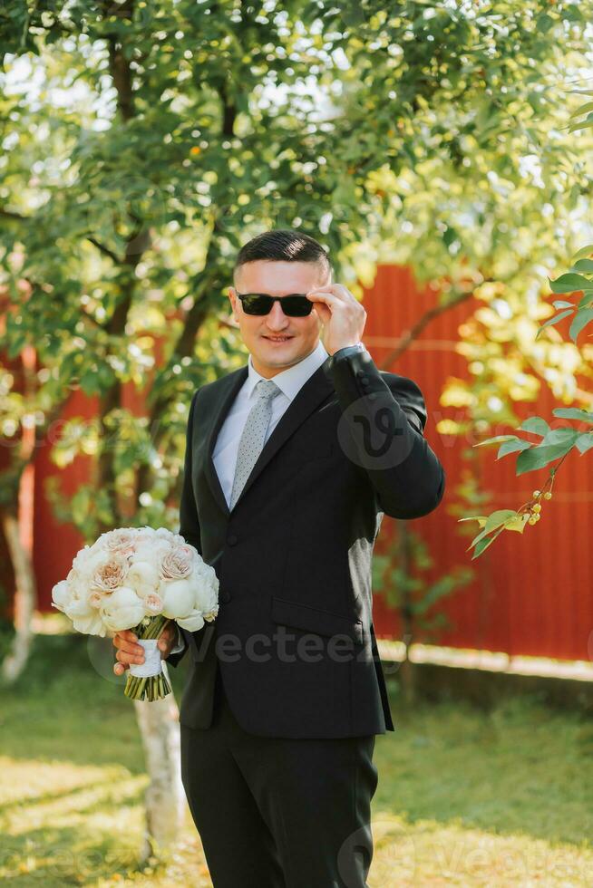 foto do a noivo dentro uma clássico Preto terno e copos, com uma ramalhete do flores dentro dele mãos