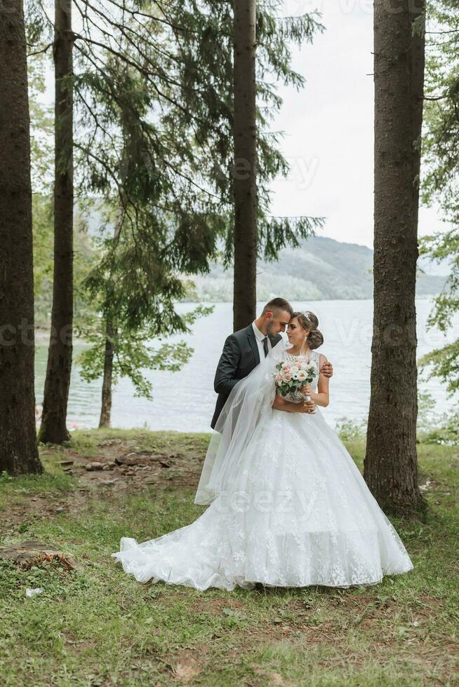 uma Casamento casal é desfrutando a melhor dia do seus vidas contra a pano de fundo do uma lago e alta árvores a noivo abraços a noiva. foto