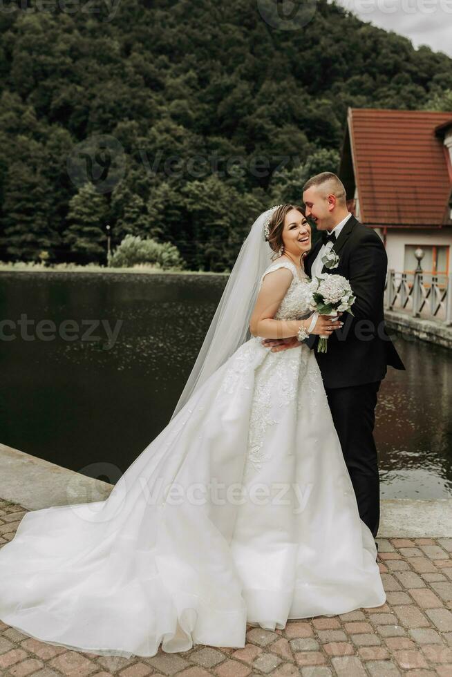 uma morena noiva dentro uma grandes vestir e uma noivo dentro uma clássico terno ficar de pé abraçando em uma ponte perto uma lago com uma castelo dentro a fundo. foto