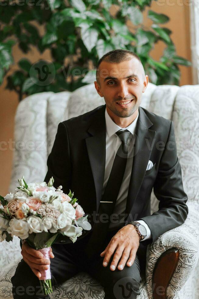 uma à moda noivo com uma ramalhete do flores é sentado em uma cadeira dentro a caro hotel sala. do noivo manhã. a noivo é obtendo pronto dentro a manhã antes a Casamento cerimônia foto