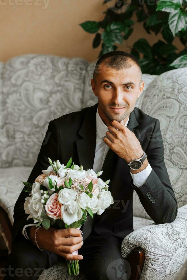 uma à moda noivo com uma ramalhete do flores é sentado em uma cadeira dentro a caro hotel sala. do noivo manhã. a noivo é obtendo pronto dentro a manhã antes a Casamento cerimônia foto