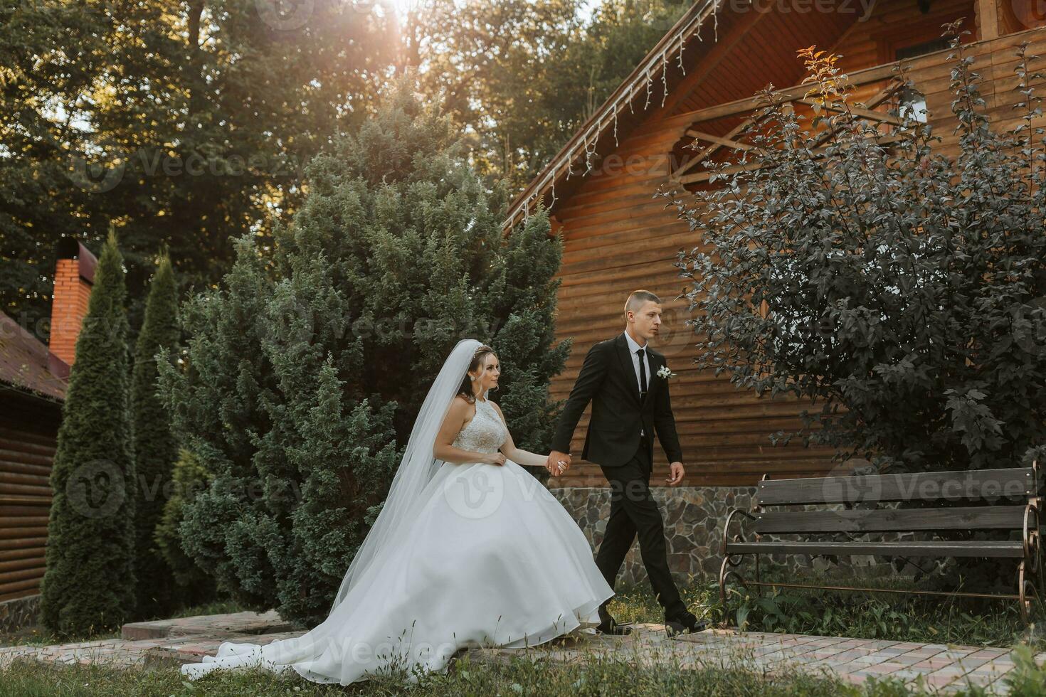 uma à moda noivo dentro uma Preto terno e uma fofa noiva dentro uma branco vestir com uma grandes véu estão abraçando e caminhando perto verde alta árvores Casamento retrato do sorridente e feliz recém-casados. foto