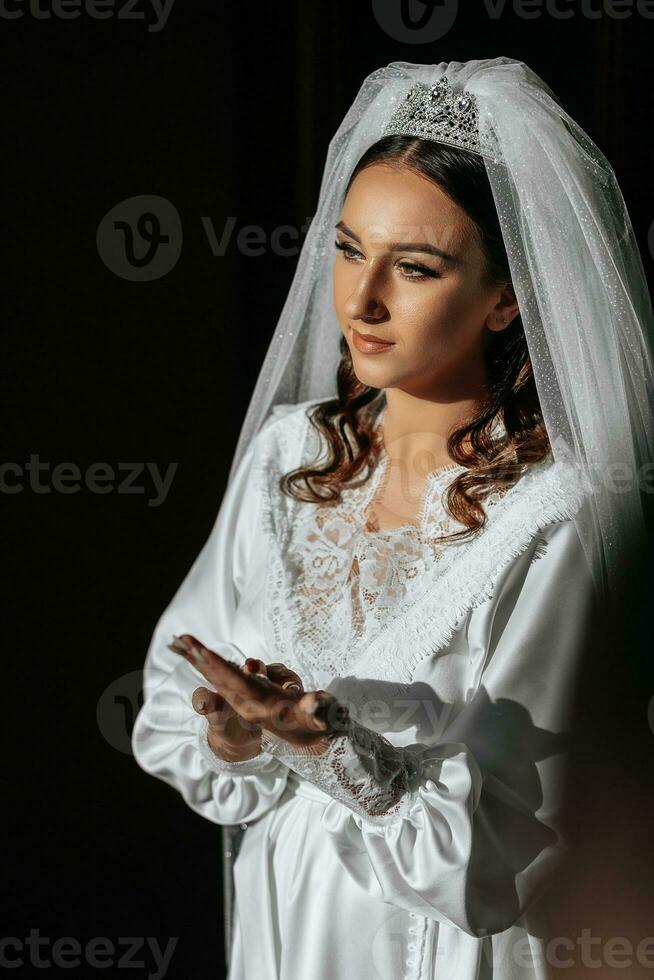 lindo noiva com grandes Sombrio encaracolado cabelo dentro uma grandes branco roupão dentro a manhã. a noiva é preparando para a casamento. profissional Maquiagem e cabelo. uma coroa em a cabeça e uma grandes véu do a noiva foto