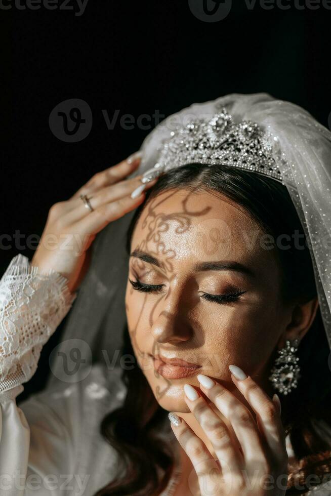 lindo noiva com grandes Sombrio encaracolado cabelo dentro uma grandes branco roupão dentro a manhã. a noiva é preparando para a casamento. profissional Maquiagem e cabelo. uma coroa em a cabeça e uma grandes véu do a noiva foto