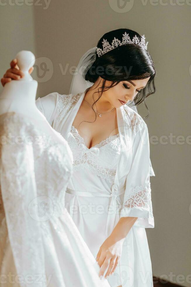 uma feliz noiva é preparando para dela luxo Casamento dentro uma hotel sala, segurando dela lindo vestir. retrato do uma mulher com profissional cabelo e Maquiagem e uma sorrir dentro uma vestir vestido foto