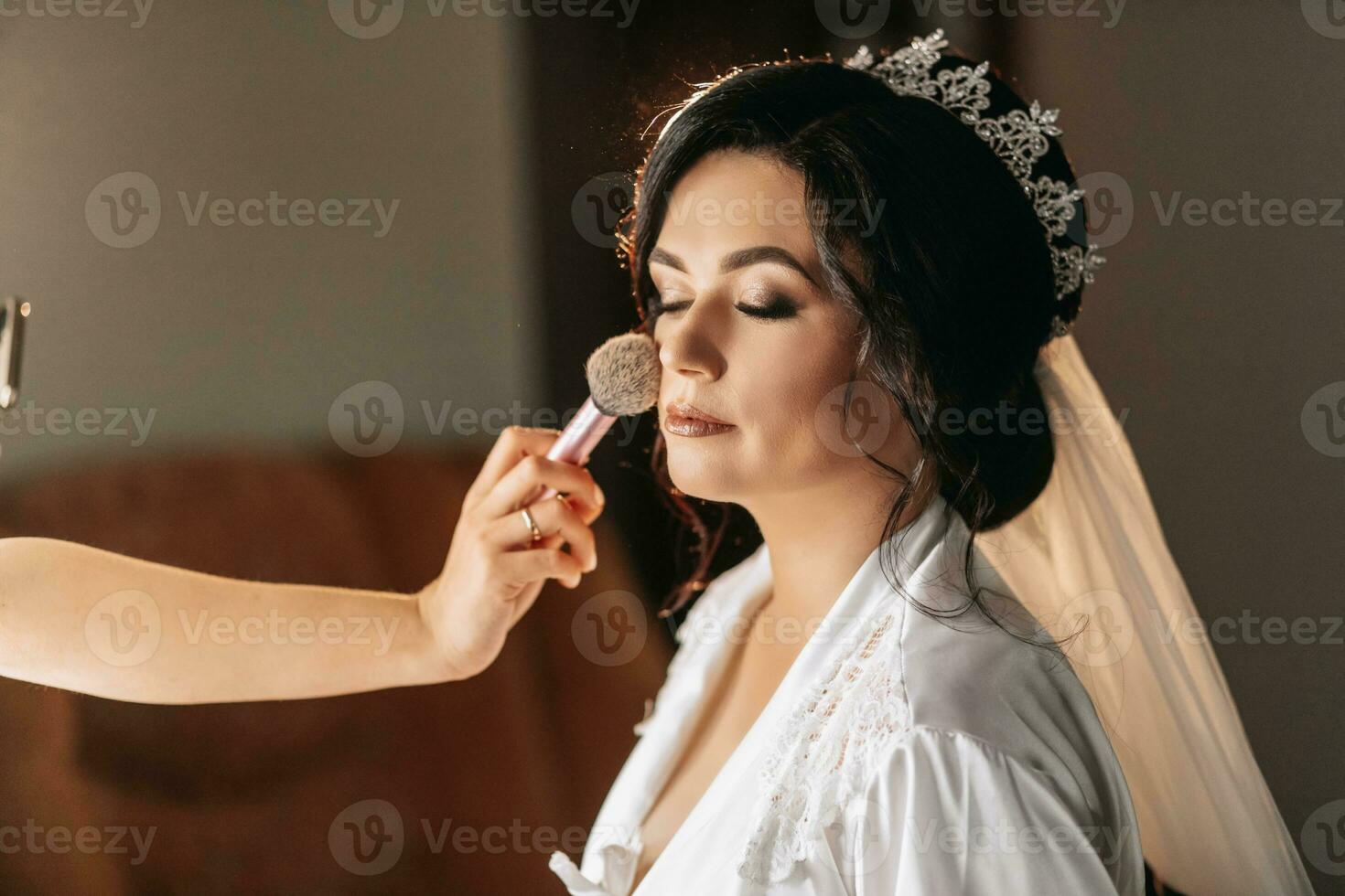 uma lindo jovem mulher com Sombrio cabelo, uma Casamento Penteado, uma coroa em dela cabeça é fazendo Maquiagem para uma Casamento ou uma foto atirar. a trabalhos do uma Maquiagem artista. manhã do a noiva. profissional Maquiagem