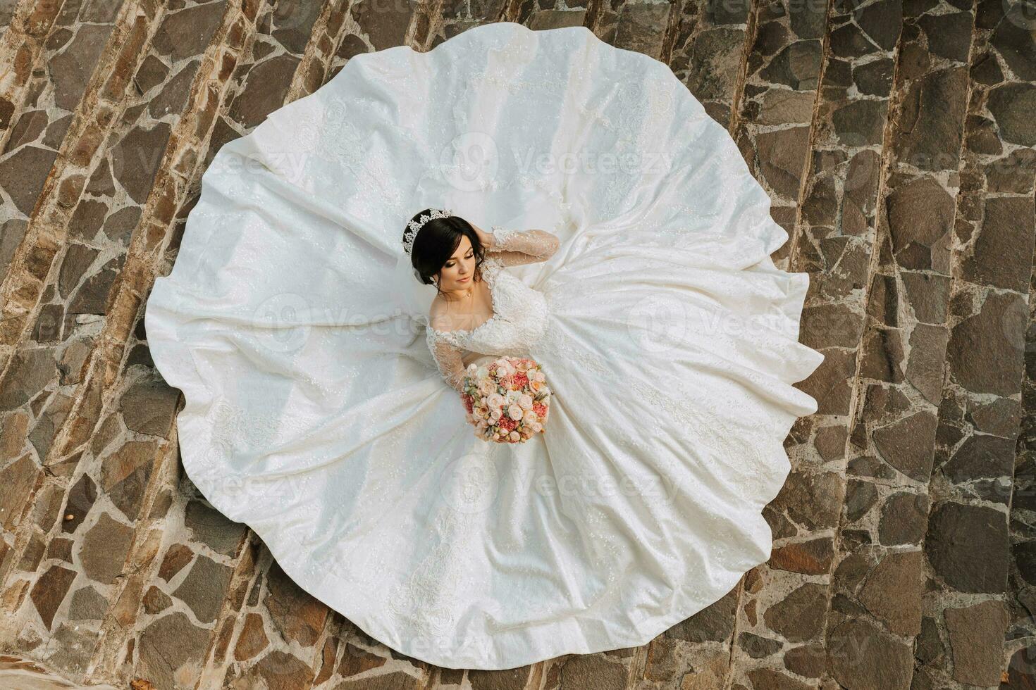 jovem lindo noiva dentro Casamento vestir e coroa em cabeça deitado em pedra passos, moda tiro debaixo severo luz solar. a foto é ocupado a partir de acima