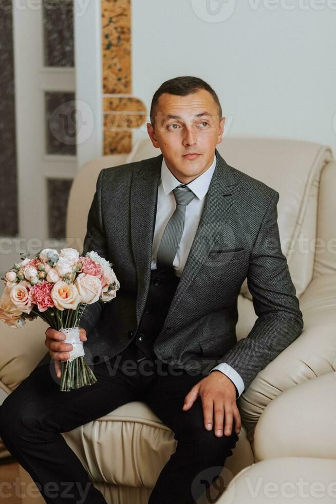 retrato. uma homem dentro uma branco camisa, uma clássico terno é sentado, posando em uma sofá, segurando uma ramalhete. uma à moda assistir. masculino estilo. moda. o negócio foto