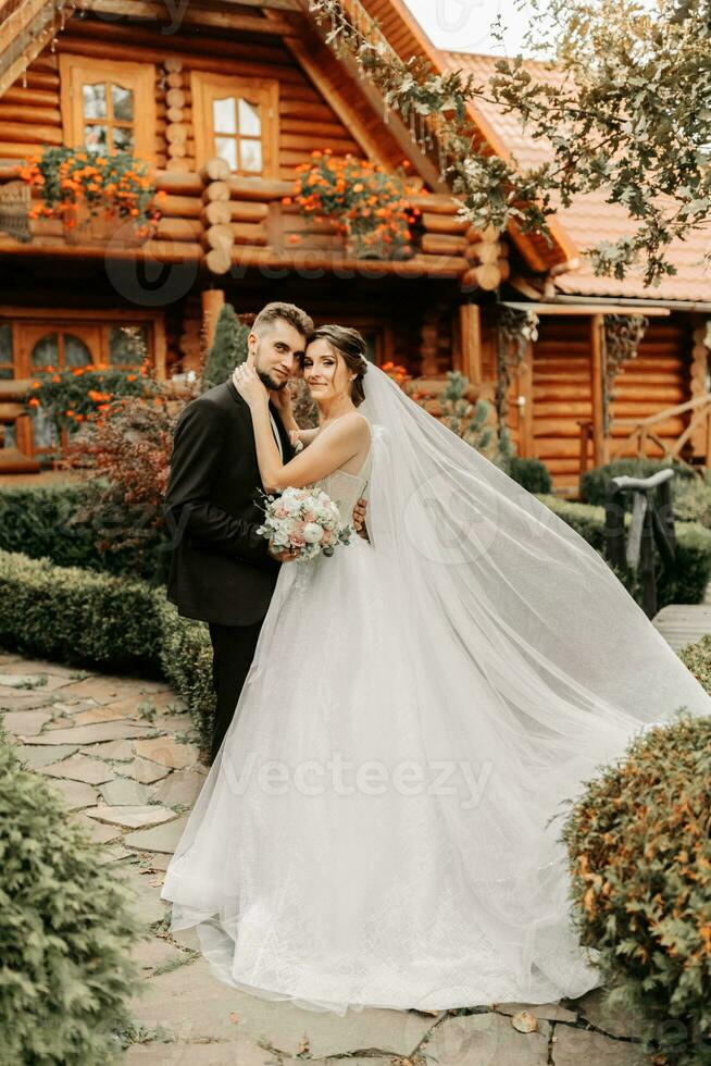 linda elegante luxuoso noiva com véu sopro dentro a vento. e uma à moda noivo beijo ao ar livre perto alta outono árvores foto