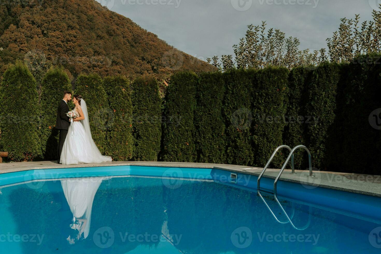 retrato do uma noiva e noivo se beijando dentro a infinidade piscina contra a céu e Alto montanhas foto