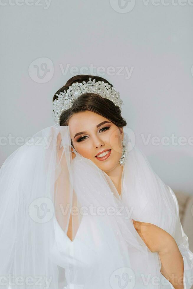 retrato do uma noiva com uma lindo Penteado e tiara em dela cabeça e natural Maquiagem posando dentro uma branco roupão dentro dela sala. a beleza do a garota. uma grandes branco véu foto