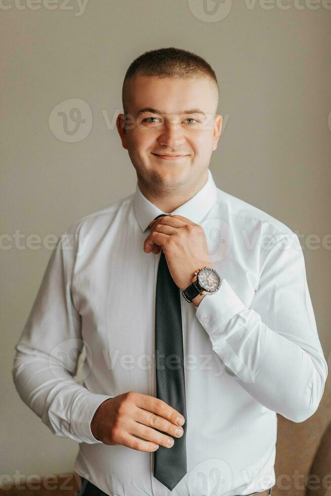 retrato do uma jovem sorridente homem dentro uma camisa dentro dele sala. a noivo pega vestido e prepara para a Casamento cerimônia foto