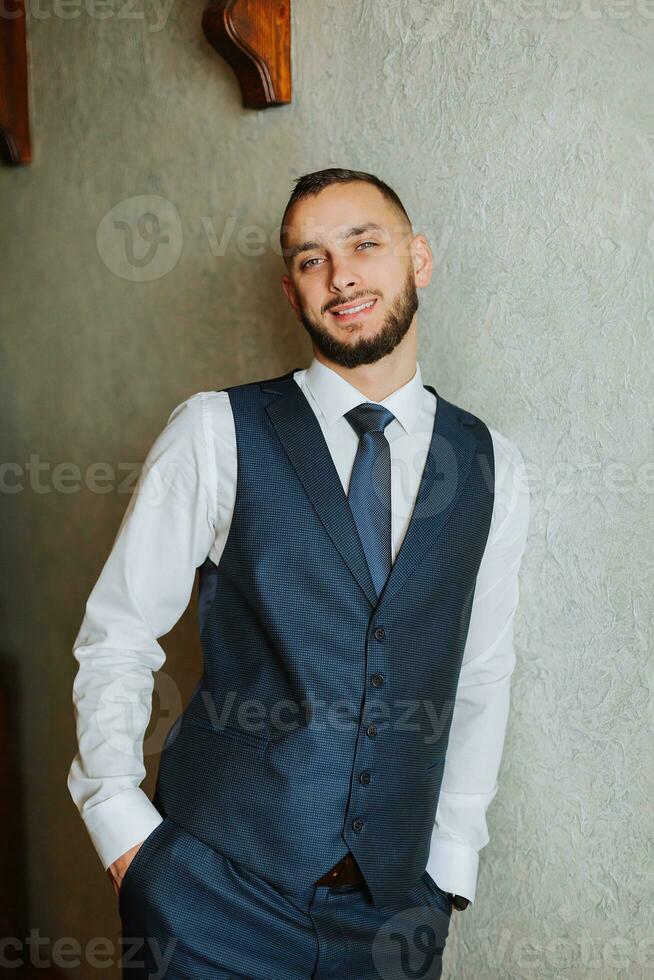 elegante noivo com uma barba dentro uma lindo azul Jaqueta e branco camisa. homem de negocios dentro clássico roupas. uma morena homem prepara para a Casamento dentro a manhã em dele Casamento dia. foto