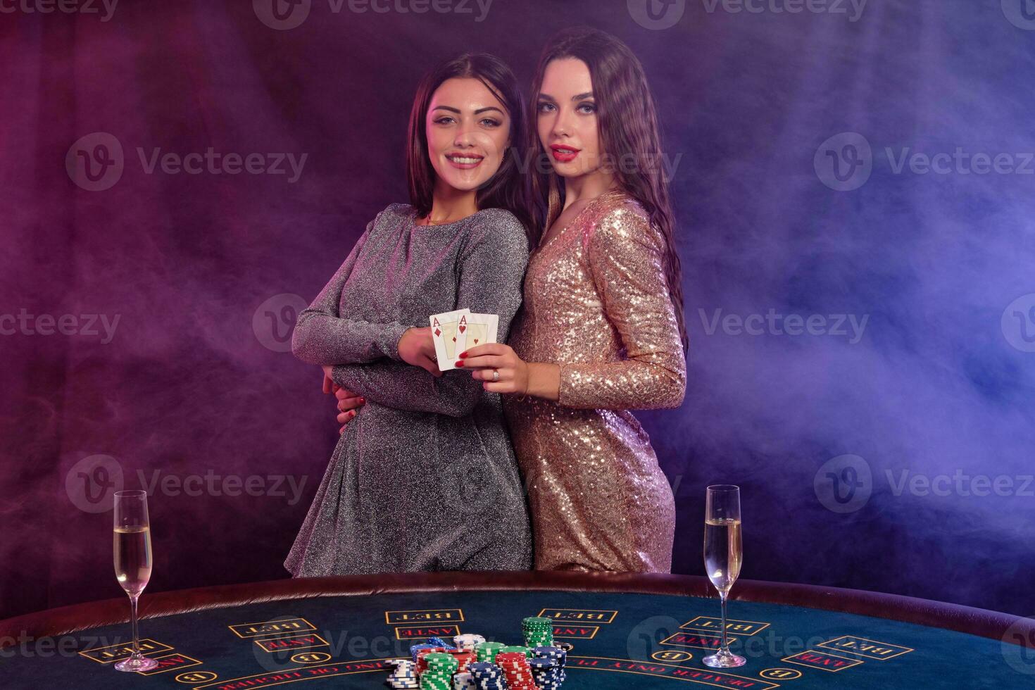dois meninas estão jogando pôquer às cassino, abraçando, segurando cartões, posando às uma mesa com salgadinhos, champanhe em isto. preto, fumaça fundo. fechar-se. foto