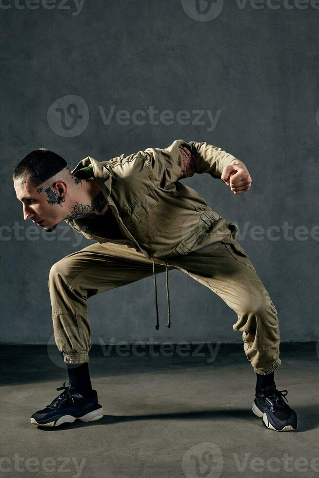 incomum homem com tatuado corpo e face, brincos, barba. vestido dentro cáqui macacão e Preto tênis. dançando em cinzento fundo. salão de dança, hip-hop foto