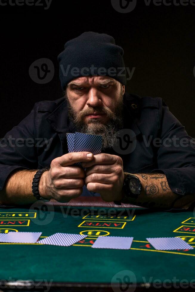 sério barbudo homem sentado às pôquer mesa e segurando cartões isolado em Preto foto