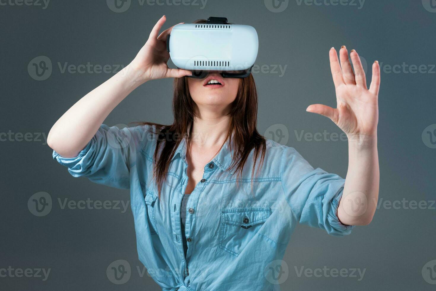 emocional jovem mulher usando uma vr fone de ouvido e experimentando virtual realidade em cinzento fundo foto