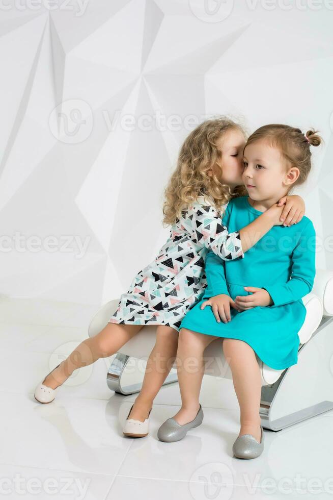 dois pequeno amigas dentro a idêntico vestidos do diferente cores sentado em uma cadeira dentro uma estúdio com branco paredes foto