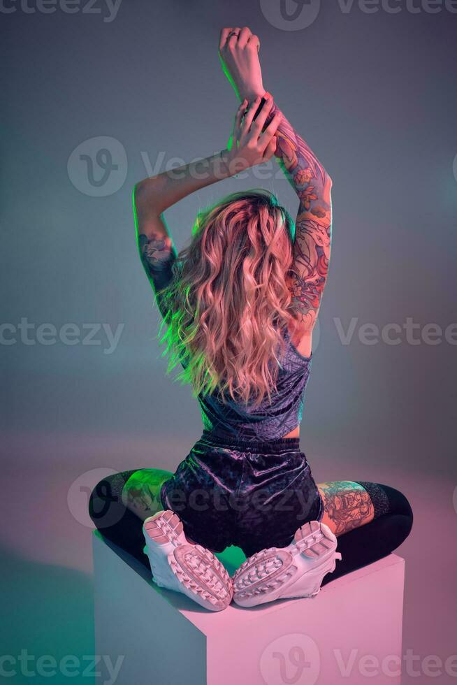 jovem ginástica Loiras mulher vestindo azul veludo saque calção posando em Câmera, senta em topo do grande cubo, costas Visão foto