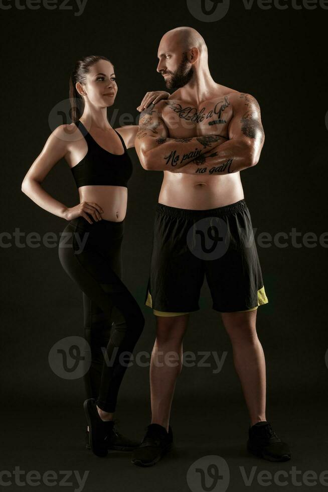 Atlético homem dentro calção e tênis com morena mulher dentro perneiras e topo posando em Preto fundo. ginástica casal, Academia conceito. foto