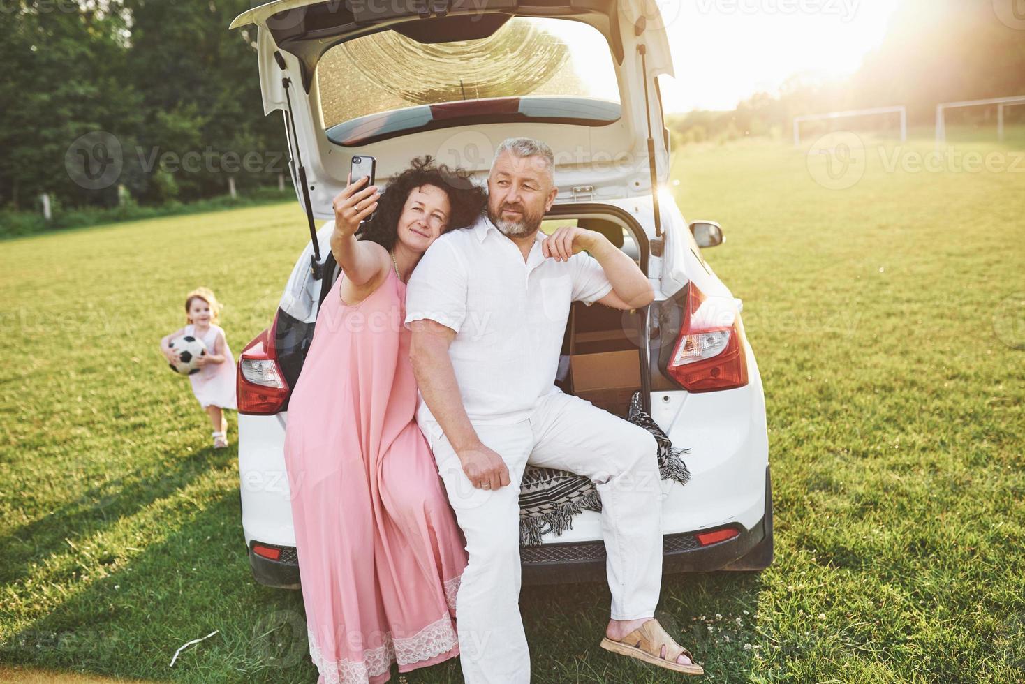 vovô e avó sentam-se juntos na natureza perto do carro. eles fazem uma selfie e a neta brinca com eles foto