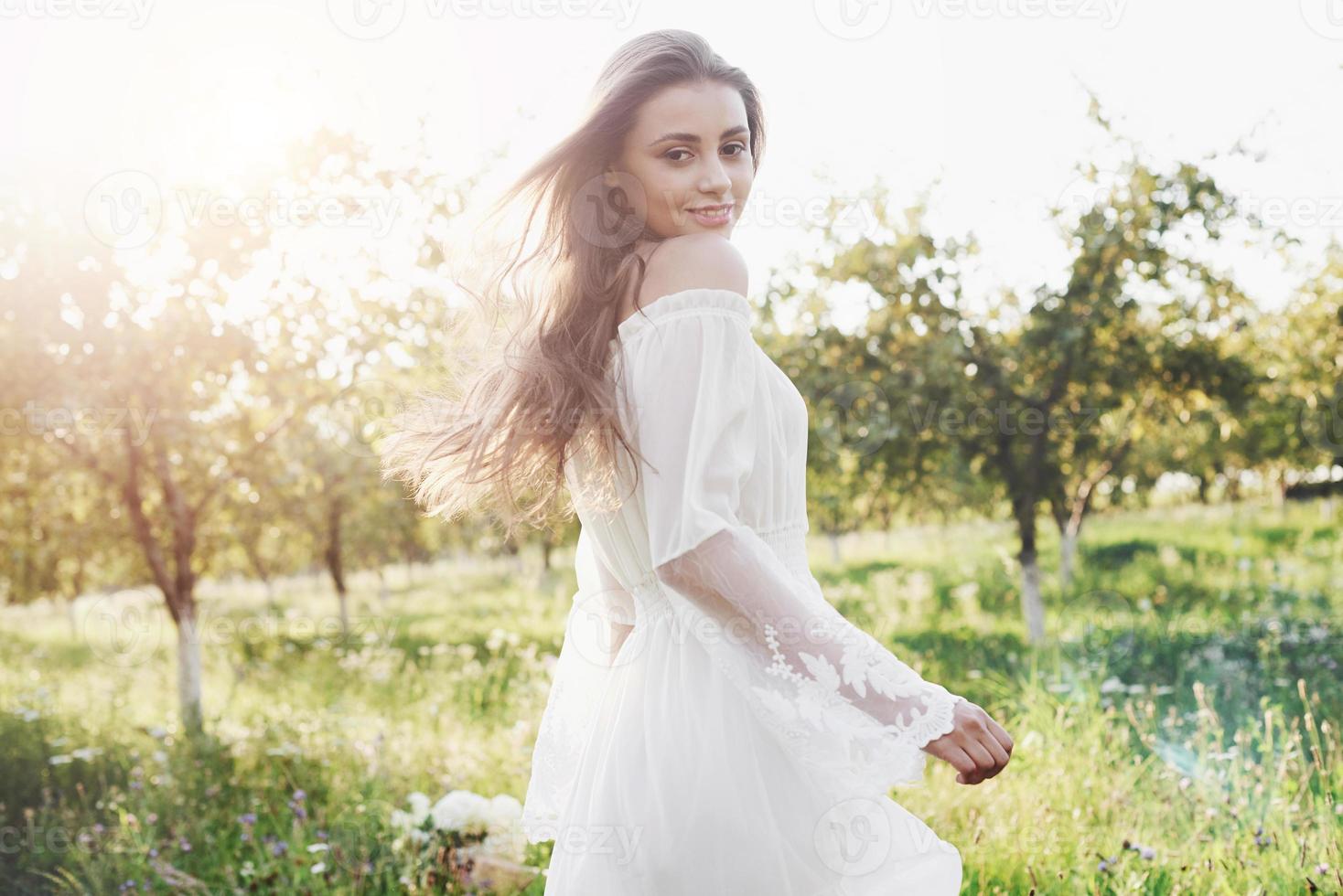 uma jovem com um vestido longo branco está caminhando no jardim. lindo pôr do sol através das folhas das árvores foto