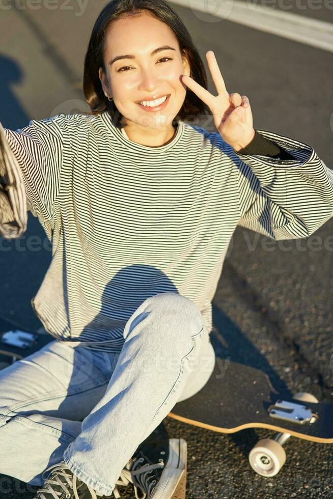 selfie do ásia menina sentado em skate, levando foto em Smartphone, sorridente e mostrando Paz sinal de v