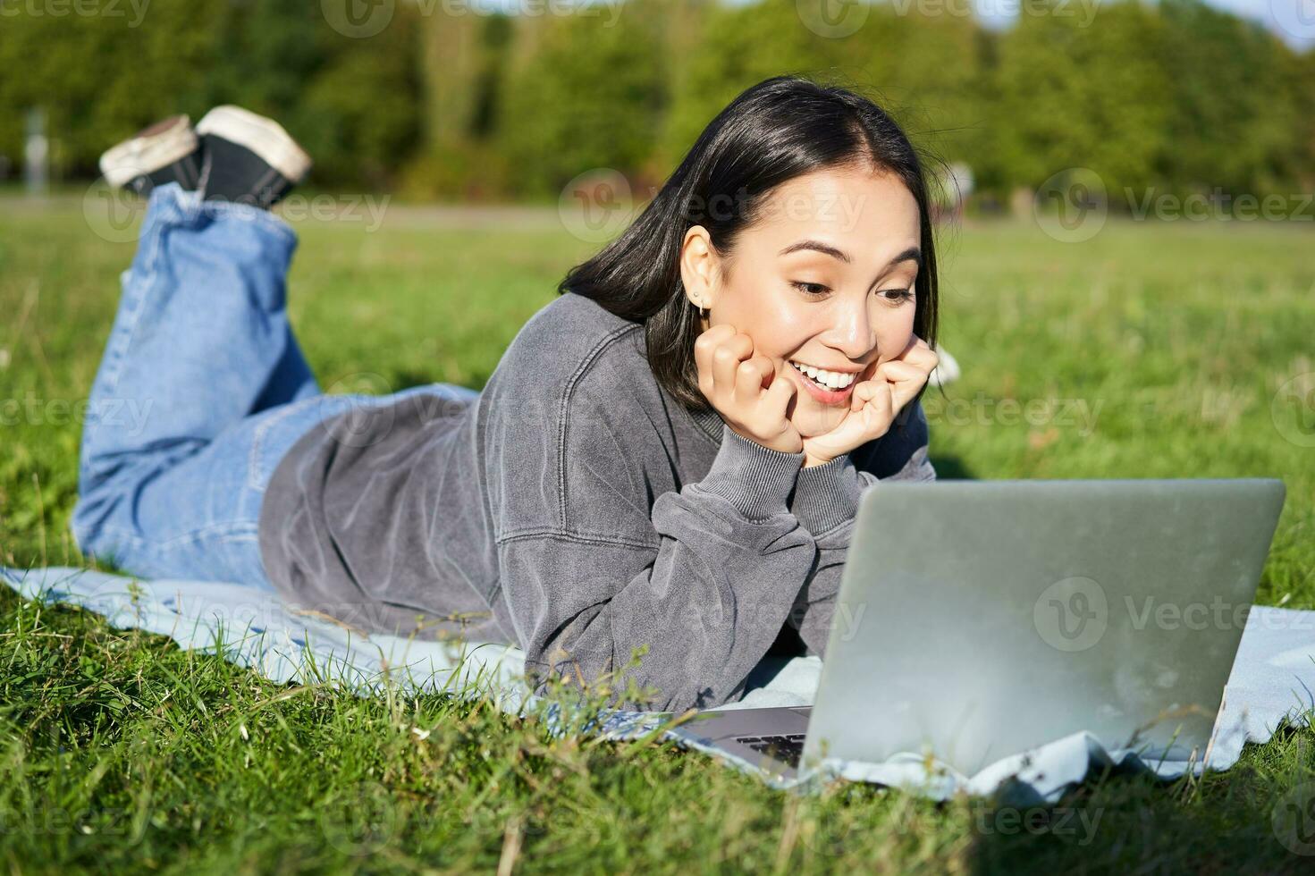 retrato do lindo menina deitado dentro parque e olhando com surpreso face às dela computador portátil, assistindo vídeos, conversando com amigos enquanto relaxante ao ar livre foto