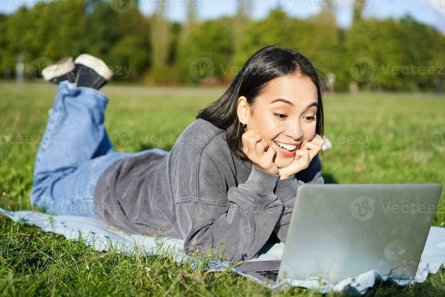 retrato do lindo menina deitado dentro parque e olhando com surpreso face às dela computador portátil, assistindo vídeos, conversando com amigos enquanto relaxante ao ar livre foto