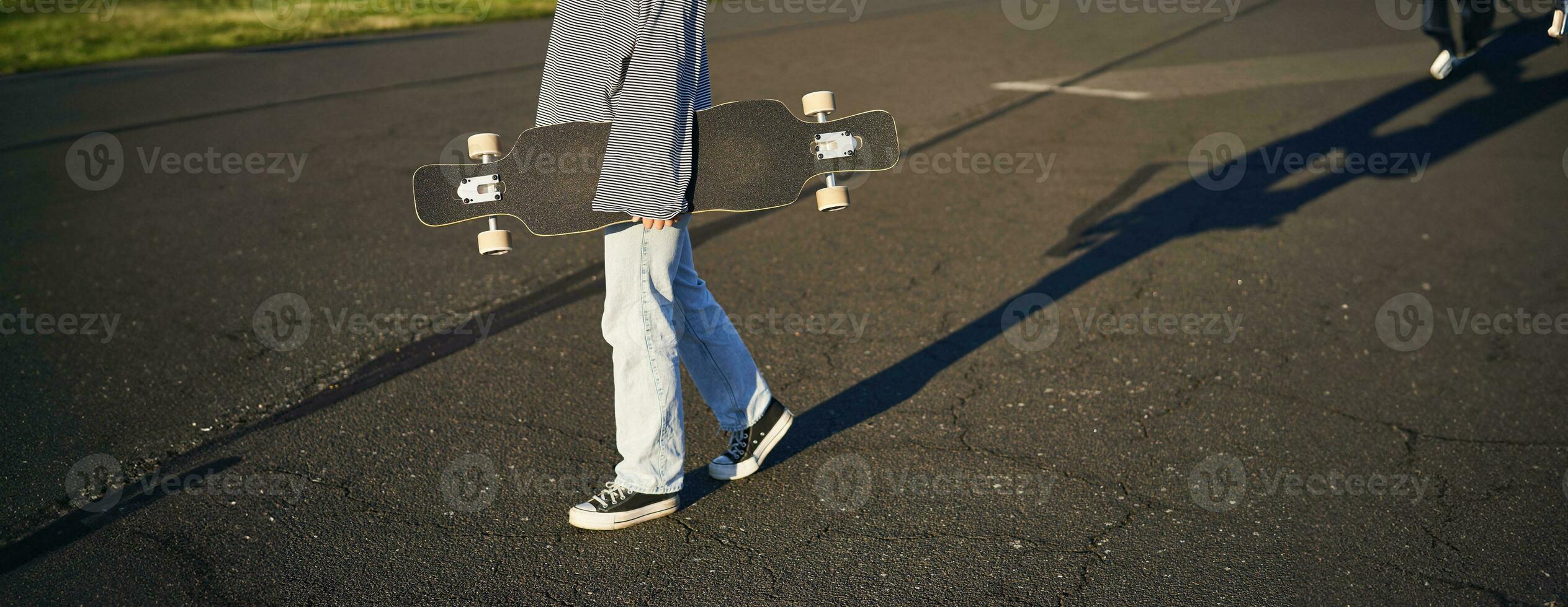 cortada tiro do adolescente menina corpo, segurando cruzador Longboard dentro mão, caminhando dentro tênis em estrada dentro jeans e suéter. jovem mulher skatista com skate foto